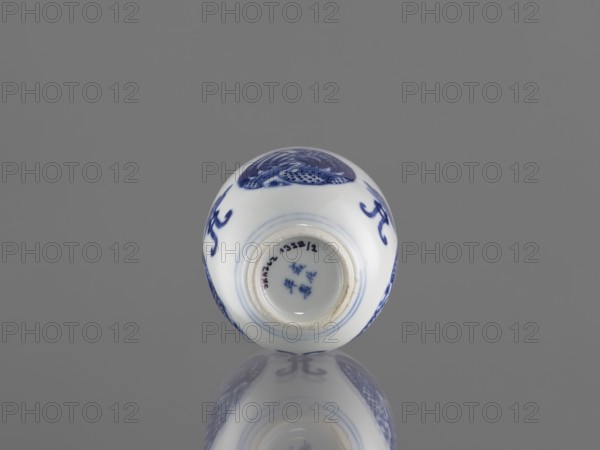 Bowl, ceramics, Chenghua 1465–1487, phoenixes, lotuses, blue-and-white porcelain, Qing style, Chinese art/culture, floral ornaments. Bowl decorated with three phoenix images and stylized lotus flowers. Early 20th century Qing dynasty, 1644–1912. China. Vessel; height 7.5 cm, diameter 8 cm., A small round ceramic object rests on a reflective surface against a solid gray background, showing white porcelain with blue painted decorative motifs and a glossy finish that creates a mirrored reflection below; the visible painted areas include curved brushstrokes and patterned designs in blue, and the rim of an opening at the top reveals the same white interior with blue detailing.