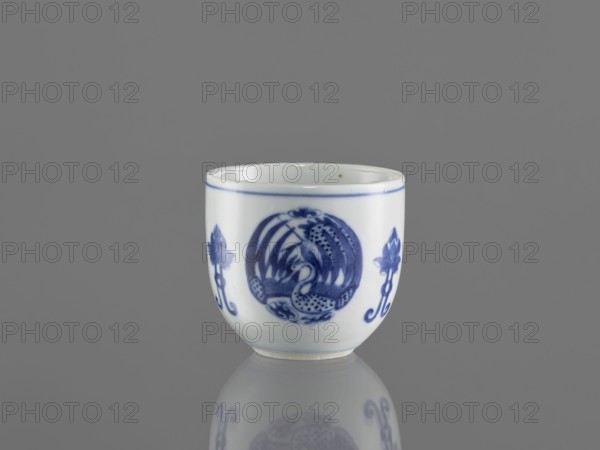 Bowl, ceramics, Chenghua 1465–1487; blue-and-white porcelain decorated with three phoenix images and stylized lotus flowers and floral ornaments; Qing style Qing dynasty, 1644–1911; early 20th century; China; vessel; height 7.5 cm, diameter 8 cm., A small white ceramic cup with blue painted decorative motifs sits centered against a smooth gray background, showing a rounded rim and a short foot; the cup features a central circular blue design and flanking floral-like blue elements, with a thin blue band near the rim and a soft reflection of the cup visible on the gray surface below.