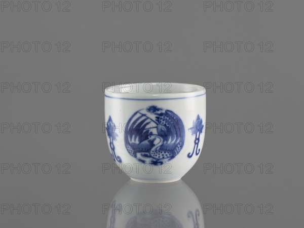 Bowl, ceramics, Chenghua 1465–1487, gift, blue-and-white porcelain, decorated with three phoenix images and stylized lotus flowers and floral ornaments, Qing style early 20th century, China, vessel; height 7.5 cm, diameter 8 cm., A small white ceramic cup sits on a reflective surface against a gray background, showing a glossy finish and a rounded form. The cup features blue painted decorations including a central circular motif with organic, curved shapes and two floral-like designs flanking it, as well as a thin blue band near the rim. The reflection of the cup and its blue designs is visible on the surface beneath it. The visible colors are white, blue, and gray.