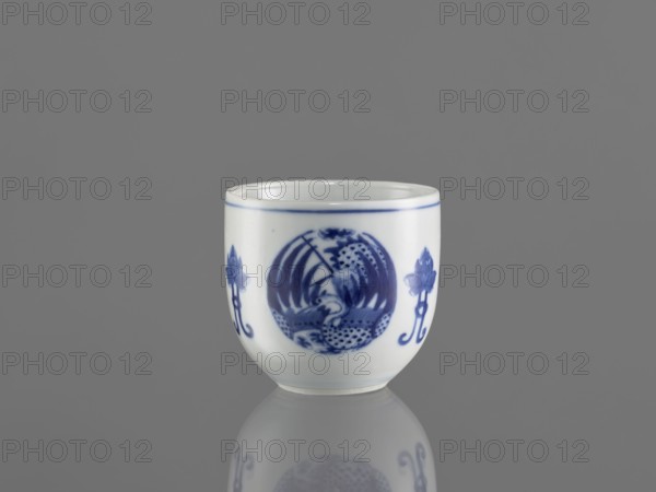 Bowl, ceramics, Chenghua 1465–1487; blue-and-white porcelain; decorated with three phoenix images and stylized lotus flowers and floral ornaments; Qing style; early 20th century Qing dynasty, 1644–1912; China; vessel; height 7.5 cm, diameter 8 cm., A small white ceramic cup with blue painted decoration sits centered on a reflective gray surface against a gray background, showing a rounded form with a thin blue rim and a central circular blue motif flanked by floral-like blue designs, with the cup's reflection visible below; visible colors include white, blue, and shades of gray.