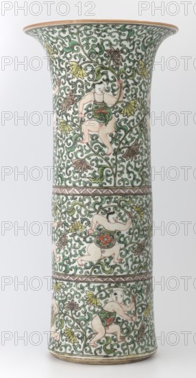 Cylindrical vase of the horn type, Chinese, Qing dynasty, Kangxi period 1662–1722, colored porcelain in the famille verte style, height 37.5 cm, diameter 18.0 cm, weight 2.35 kg; decorated with lotus tendril ornament and belt decoration., A tall flared ceramic vase with an off-white base color is covered in an all-over scrolling green vine pattern with stylized leaves in green, yellow, and brown; the surface is decorated with a group of playful cherubic figures with pale skin and dark hair wearing small green garments adorned with red and orange floral motifs, shown in various running and climbing poses among the vines; thin brown decorative bands encircle the vase separating the sections of the design; the rim has a warm beige edge and the foot shows a slightly darker beige ring.