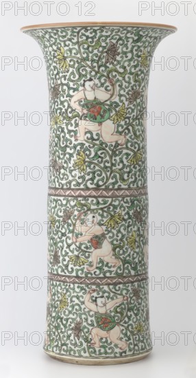 Cylindrical vase of the horn type, Chinese, Qing dynasty Kangxi period, 1662–1722, colored porcelain in the famille verte green family palette, decorated with lotus tendril ornament and belt decoration in the famille verte style; ceramics vessel, height 37.5 cm, diameter 18.0 cm, weight 2.35 kg., A tall flared ceramic vase decorated with an all-over scrolling vine pattern in green and outlined in dark brown, featuring a group of small stylized human figures with pale skin tones positioned among the foliage; the figures wear green and red garments and are shown in various playful poses. The vine design includes several stylized leaves and blossoms in shades of yellow and muted brown set against a white background. The vase has a narrow midsection separated by decorative bands in brown and the rim and base have thin brownish-gold accents. The overall color palette includes white, green, brown, yellow, red, and pale skin tones.