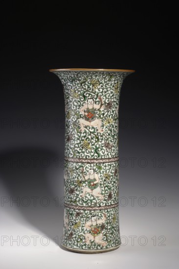 Cylindrical horn vase, Chinese, Qing dynasty, Kangxi period 1662–1722, colored porcelain in the famille verte palette. Ceramic vessel decorated with lotus tendril ornament and belt decoration. Height 37.5 cm; diameter 18.0 cm; weight 2.35 kg., A tall flared ceramic vase standing on a smooth surface, decorated all over with an intricate scrolling vine pattern in green, set against a white background with accents of brown, yellow, orange, and pale flesh tones; the surface shows a repeating motif of three seated figures positioned within the vinework, each figure wearing a green garment and holding a round orange object, and the vase is divided visually by a few narrow horizontal decorative bands in brown.