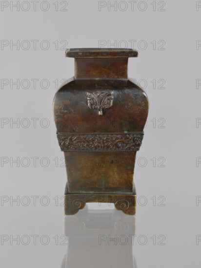 Fanggu vessel, Chinese, Qing dynasty 1644–1911, 19th century. Ceremonial vessel depicting dragons and fantastical beasts, with meander ornament and backdated signatures. Material/technique unspecified. Dimensions: height 13.4 cm, width 9.0 cm, depth 7.9 cm., An upright rectangular metal vessel with a dark brown and greenish patina stands on a glossy surface, showing its reflection beneath; the vessel has a narrow flat top, a slightly curved upper body, a band of raised ornate relief around its middle, and a straight lower section that rests on a small base with scroll-shaped cutouts; a decorative raised emblem appears on the curved upper face, and the metal shows mottled areas of darker brown, bronze, and hints of green.