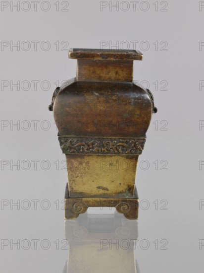 Fanggu vessel, Chinese, fanggu vessel, dragons iconography, Chinese art, ceremonial vessel, Qing 1644–1911, beasts fantasy, meander ornament, backdated signatures, 19th century, China, vessel; height 13.4 cm, width 9.0 cm, depth 7.9 cm., An upright rectangular metal vessel with a lid and a stepped top, showing a patina of brown, dark bronze, and hints of green, with a band of ornate raised decoration around its middle, a slightly curved upper body, a straighter lower section in a lighter brass tone, and a pair of scroll-shaped feet at the base; the piece sits on a reflective surface that mirrors its colors.