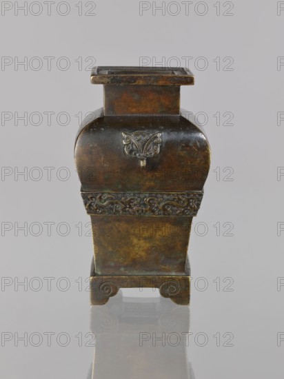 Fanggu vessel, Chinese, fanggu vessel, dragons iconography, Chinese art, ceremonial vessel, Qing 1644–1911, beasts fantasy, meander ornament, backdated signatures, 19th century, China, vessel, height 13.4 cm, width 9.0 cm, depth 7.9 cm, A small metal container with a square opening and a rectangular neck sits upright; its body has a curved upper section and a tapered lower section, with decorative raised relief on the front and a band of ornate carved pattern around the middle. The piece rests on a short base with curled scroll-like feet and is reflected faintly on the glossy surface beneath it. The visible colors include dark brown, greenish patina, golden bronze highlights, and subtle blackened areas, all set against a plain light gray background.