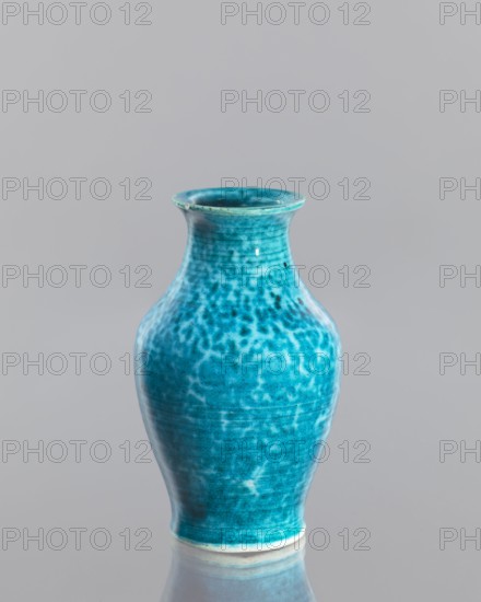 Vase, maker, Chinese, Qing dynasty 1644–1911, 2nd half of the 18th century, glazed crackleware ceramic vessel, height 12.5 cm, diameter 7.6 cm., A turquoise ceramic vase with a flared rim and a rounded body sits against a smooth light gray background, its surface showing mottled darker and lighter turquoise patterns and a glossy finish that produces subtle highlights and reflections, and a pale unglazed band is visible at the base where the vase meets a reflective surface that mirrors its shape.