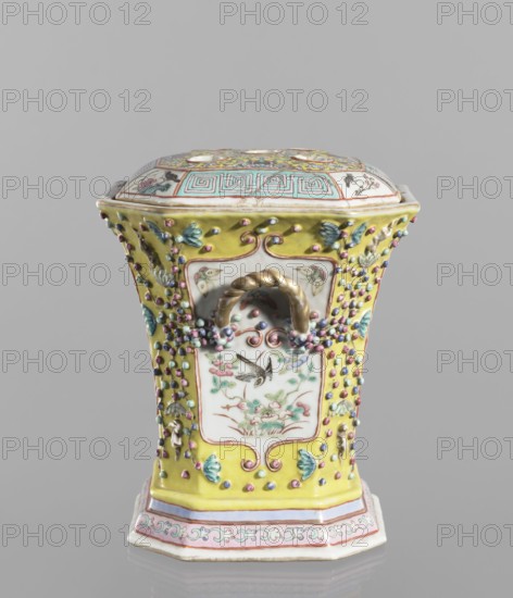 Vase with a lid; ceramics, colored porcelain; openwork, flat perforated lid; decorated with flowers, birds, plant ornaments, meander and lotus tendril motifs; 19th century, China; vessel; height 27.0 cm, width 15.0 cm., An ornate lidded ceramic vessel with a hexagonal flared shape, decorated in bright yellow, white, pink, turquoise, green, blue, gold, and lavender. The lid has patterned panels with turquoise geometric motifs and floral designs in yellow and pink, and the body features white inset panels with painted botanical and bird scenes surrounded by raised bead-like appliqués in pink, blue, and purple. There are small molded leaf and bud decorations in green and turquoise, a twisted gold-colored handle on the front, and a stepped base banded in pink, white, and turquoise with scrolling floral ornament. The surface shows a glossy glaze and the object is set against a plain gray background.