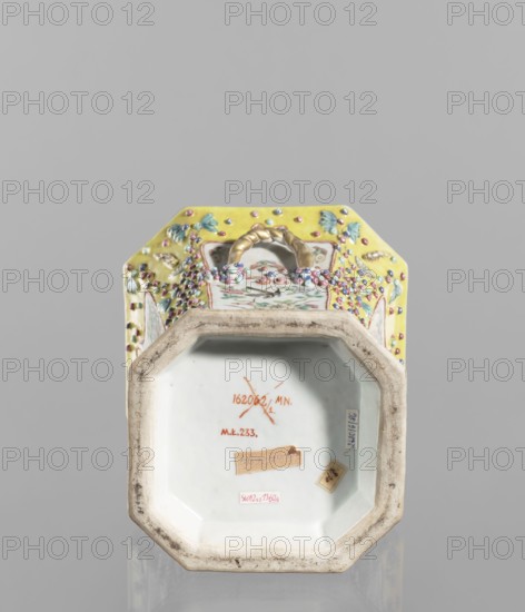 Vase with a lid, ceramics, colored porcelain, openwork perforated flat lid, decorated with flowers, birds, meander ornament, plant ornaments, lotus tendril ornament; 19th century, China; vessel; height 27.0 cm, width 15.0 cm., An upside-down octagonal ceramic object with a decorative exterior in yellow, pink, green, blue, white, gold, and purple tones; the visible top edge shows a braided gold-colored handle and raised beaded embellishments in pink and purple over painted floral and butterfly motifs in green and blue on a yellow ground; the exposed base interior is white with a worn beige rim and several small paper labels affixed; the background is a uniform light gray.