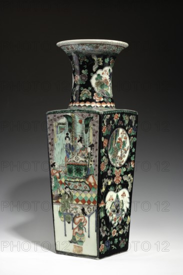 Vase from the famille noire, colored porcelain, painted decoration in medallions featuring butterflies, plant motifs, and figural scenes; Qing dynasty, Kangxi period 1662–1722; Chinese ceramics; vessel, height 44.5 cm, width 14.5 cm, depth 14.3 cm., A tall hexagonal porcelain vase with a flared rim sits against a dark background, showing a glossy surface decorated in a palette of black, white, green, red, pink, blue, and gold tones; one face displays a painted scene of several figures in traditional dress around a table with patterned flooring and drapery, while adjacent faces feature floral motifs and circular cartouches containing stylized flowers and leaves; the neck is adorned with floral and cloud-like patterns on a black ground, and the rim is white with a pale decorative band beneath it; the vase casts a soft shadow on the pale surface beneath.