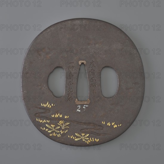 Crossguard tsuba, Ichiryûsai Masamitsu; Shôami school. Oval tsuba tatemaru gata decorated in high relief with colored metals iro-e zôgan: one side shows a stream winding among grasses and dandelions; the other depicts a buffalo and a she-buffalo grazing in a flowery meadow. Edo period, 1st half of the 19th century Japan. Materials: metals with colored inlay. Dimensions: height 7.6 cm, width 7.6 cm, depth 0.4 cm., A round, dark brown metal disc with a smooth matte surface and a central elongated cutout flanked by two oval cutouts, showing areas of subtle rust and wear. Near the lower edge there is a winding engraved line suggesting a stream, decorated with a few small gold-colored leaf or grass motifs and several gold-colored floral or plant elements. The metal around the central cutout shows lighter worn patches in a beige tone, and there are faint darker and lighter variations across the surface. The visible colors are dark brown, gold, beige, and gray.