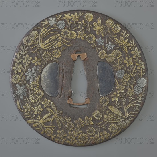 Crossguard tsuba, Mino school, Japanese, Edo period 1603–1868, 18th century. Round tsuba maru gata with rounded edge maru mimi, ryohitsu ana holes plugged with shakudô alloy, copper sekigane. Decorated on both sides with dense, low-relief inlay of brass and silver forming an ornament of various flowers, including praying mantises, butterflies and dragonflies. Signed. Diameter 8.3 cm; thickness 0.3 cm., A round metal object with a dark brown surface decorated with a floral and insect motif in gold and silver tones, featuring a central elongated keyhole-shaped opening and a few irregular metal fittings in a copper color, with two oval dark gray insets flanking the opening and a background of gray around the object.