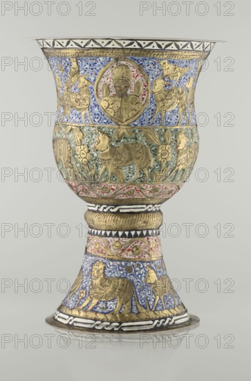 Cup, Persian, Qajar dynasty 1785–1925, 19th century; vessel, height 25.5 cm, diameter 18.4 cm. Decoration: cartouches with a bust of a man, animals lions, birds, figures, figurative motifs, flowers, geometric and small plant ornament., A decorated metal goblet with a flared bowl and a flared foot standing on a shallow circular base, covered in ornate relief figures and vegetal patterns. The surface shows gilded raised figures of animals and humanlike faces set against painted backgrounds in blue, green, and red, with areas of white and black geometric bands. The upper bowl has a band of blue background with gilded animals and a central gilded humanlike portrait surrounded by painted accents, the midsection features a greenish background with gilded animals and floral motifs, and a band of red and white painted decoration separates the bowl from the stem. The stem has alternating bands including a gilded ribbed section and a black and white patterned band, while the foot is decorated wi...