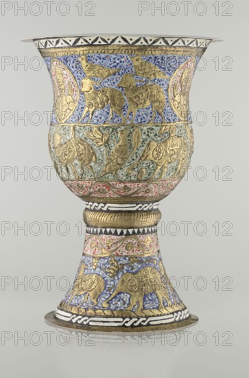 Cup, Persian, Qajar dynasty 1785–1925, 19th century. Ceramic vessel, height 25.5 cm, diameter 18.4 cm. Decoration: cartouches with a bust of a man, animals lions, birds, figures, flowers, geometric and small plant ornament; figurative motifs., A decorative goblet-shaped metal vessel with a flared rim and a pedestal base, covered in ornate embossed and painted designs; the primary colors visible are gold, blue, green, red, black, and white. The upper bowl area shows gold animal figures against a blue patterned background, with small circular medallion shapes at the sides containing stylized human faces in gold. Below that, a band features gold birds and other animals on a green leafy background, followed by a red and white decorative border. The narrow central stem has a ribbed gold section and patterned white and black bands, while the lower flared foot is decorated with gold elephant figures set on blue patterned panels and additional red and gold scrollwork, all sitting on a circular...