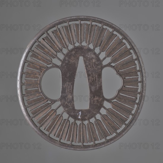 Crossguard tsuba, Tsunetada maker; decorated with an openwork positive motif of thirty-two concentrically arranged arrow shafts and feathers with fine engraved details; round tsuba maru gata with a rounded edge maru mimi. Inscription/association: Bushu Musashi. 19th century Edo period, 1603–1868. Japan, Kawagoe. Materials/typology: tsuba sword crossguard, weapons/swords katana. Dimensions: height 7.2 cm, width 7.0 cm, depth 0.3 cm., A round metallic object with a central elongated oval opening and two rounded openings on either side, featuring a decorative radiating pattern of narrow, feather-like segments around the inner perimeter; the metal shows a dark brown and gray patina with areas of lighter brown highlights and subtle reddish-brown tones, set against a neutral gray background.