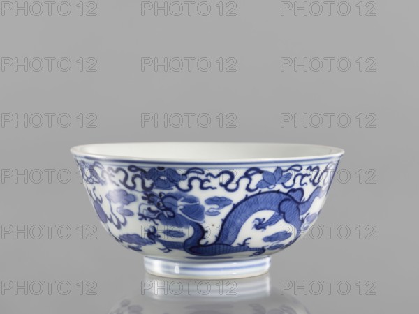 Bowl, ceramics with pearls, blue and white porcelain in Qianlong/Qing style, dragons iconography; At the top of the bowl an ornamental band with eight Buddhist symbols. Date: 1736–1795 Qianlong period, China; vessel; height 5.5 cm, diameter 12.4 cm., A porcelain bowl sits on a reflective surface against a neutral gray background, showing a rounded form with a flared rim and a short foot. The bowl's exterior is white with intricate blue painted decoration featuring sinuous scrolling motifs and a stylized creature with a scaled body incorporated into the pattern. The blue decoration varies in tone from deep blue to lighter blue washes, and a thin blue band circles the rim and the foot. The interior rim appears plain white, and the reflection beneath the bowl shows the white and blue colors mirrored on the surface.