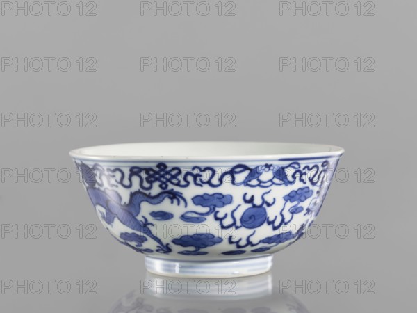 Bowl, ceramics with pearls, blue and white porcelain in Qianlong/Qing style, decorated with dragons; at the top of the bowl an ornamental band with eight Buddhist symbols. Chinese, Qianlong period 1736–1795 / Qing dynasty 1644–1911. Vessel; height 5.5 cm, diameter 12.4 cm., A porcelain bowl with a white base and blue decorative painting around its exterior, showing a pattern of stylized motifs and cloud-like shapes; the rim has a thin blue band, and the bowl sits on a short foot with subtle blue ringing; the background is a uniform light gray and the surface beneath the bowl reflects a soft, muted reflection.
