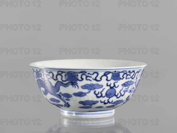 Bowl, ceramics and pearls, blue-and-white porcelain in Qianlong/Qing style, 1736–1795 Qianlong period, China. Vessel with dragons iconography; at the top of the bowl an ornamental band with eight Buddhist symbols. Height 5.5 cm; diameter 12.4 cm., A shallow ceramic bowl with a white interior and exterior decorated with blue painted motifs, including flowing cloud-like shapes and a stylized dragon form, sitting on a short foot and reflected faintly on a glossy surface, against a plain light gray background; visible colors include white, several shades of blue, and light gray.