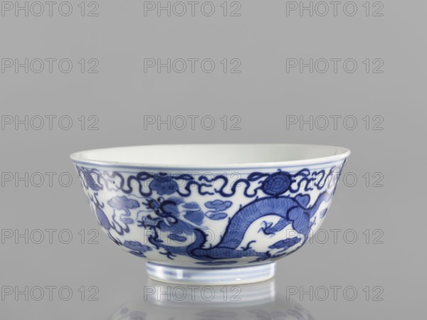 Bowl, ceramics with pearls, blue-and-white porcelain in Qianlong/Qing style, decorated with dragons and an ornamental band with eight Buddhist symbols at the top; Chinese, Qing dynasty Qianlong period, 1736–1795; vessel; height 5.5 cm, diameter 12.4 cm., A shallow ceramic bowl sits on a reflective surface against a plain gray background, showing a white interior and exterior decorated with a flowing blue painted design that includes a sinuous dragon motif, stylized cloud-like shapes, and ornamental borders; the rim and foot of the bowl have subtle blue accents, and the overall visible colors are white, various shades of blue, and gray.