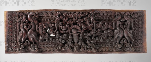 Decorative panel;, wooden panel likely originally a decorative element, part of home furniture or a door. The scene shows five pairs of birds—smaller parrots Skt. suka and larger geese Skt. hamsa—surrounded by lush plants, with a background of abundant floral and geometric ornament. Indian, South Asian; Indian art; birds; floral ornament; flowers; tree iconography. Sculpture, 18th century; height 24.0 cm, width 77.0 cm., A rectangular carved wooden panel with a dark brown to reddish-brown surface showing intricate raised relief work across its length; a pair of stylized bird-like creatures with curved necks and patterned bodies appear at each end, facing inward, and are flanked and connected by swirling vegetal scrolls and floral motifs; the central area features a clustered arrangement of carved rosettes and rounded blossom forms above a vase-like element, all surrounded by ornate lattice and repeated geometric border patterns; the wood shows lighter brown edges and small areas of wea...