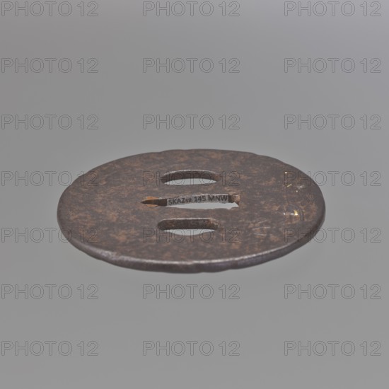 Crossguard tsuba, Kaneie craftsman / school, 18th century Edo Period, 1603–1868, Japan; Fushimi, Yamashiro province. Oval tsuba tatemaru gata, rounded edge maru mimi; height 8.0 cm, width 7.4 cm, depth 0.4 cm. On both sides decorated in relief with landscape scenes and figures: obverse — a wanderer with two calabashes at his belt and a stick standing by a mountain stream; some details face, hands, bottles, binding inlaid with silver, gold, and copper; reverse — a fisherman in a boat by a bamboo-covered shore with minor accents inlaid with gold. Themes: landscape motifs, rocks, trees, bamboo, water, fishermen, boats, sea waves; function: sword crossguard katana tsuba., A flat round metal disc with a weathered, rusty brown surface and a slightly irregular edge, featuring a few elongated oval cutouts and a narrow horizontal slot; the metal shows patches of darker brown and faint hints of gold, and the background is a uniform soft gray.
