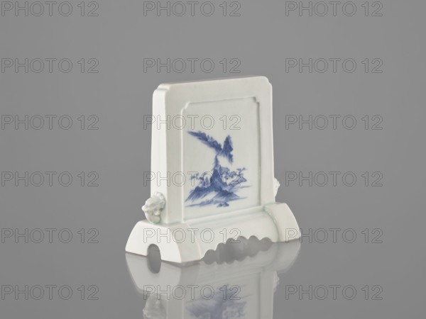 Decorative screen, ceramics; blue-and-white porcelain; Chinese, Qing dynasty 1644–1911; 19th century. Base cut with a wavy undercut; mascarons on the sides. Mountain landscapes depicted on both sides of the screen. Height 13.5 cm, width 13.7 cm., A small ceramic object stands on a reflective surface, predominantly white with blue painted decoration on its front panel; the piece has a rectangular upright section with a framed recessed panel showing a blue landscape motif, flanked by a pair of sculpted protrusions on the sides, and it rests on a curved base with an arched cutout and scalloped edge. The visible colors include white, blue, gray from the background, and subtle greenish tints on the glaze.