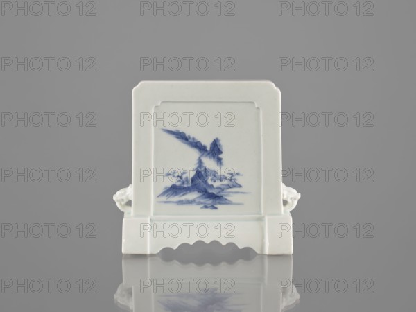 Decorative screen, ceramics; blue-and-white porcelain; Qing dynasty 1644–1911; 19th century, China. Base cut with a wavy undercut; mascara on the sides. Mountain landscapes painted on both sides of the screen. Height 13.5 cm, width 13.7 cm., A small ceramic screen-like object stands upright on a reflective surface, showing a central recessed panel with a blue-painted landscape scene. The piece is primarily white with blue decoration, and it has a rectangular frame with rounded inner corners and a scalloped base edge. A pair of small lion-like handles protrude from the sides, and the object casts a soft reflection below. The background is a uniform gray.