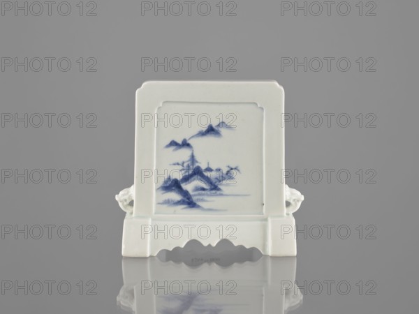 Decorative screen, ceramics, blue-and-white porcelain, Chinese, Qing 1644–1911, 19th century. Base cut with a wavy undercut; mascara on the sides. Mountain landscapes depicted on both sides of the screen. Height 13.5 cm, width 13.7 cm., A small rectangular ceramic object with a pale off-white glaze stands on a flat reflective surface, its front face inset with a framed panel painted in a muted blue depicting a stylized landscape of mountains and water; the piece has a stepped base with scalloped cutouts and small sculpted side handles that extend outward, and the overall colors visible are off-white, blue, gray from the background, and subtle darker shadows in the reflection.