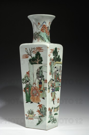 Vase, Chinese, Qing dynasty Kangxi period, 1662–1722. Porcelain, colored green family. Cylindrical-neck vase, height 50.0 cm, width 14.8 cm, depth 14.8 cm. Walls decorated with figural scenes depicting horsemen, courtiers, and women; symbolic ba bao emblems on the collar; mountain landscape with staffage on the neck., A tall, square ceramic vase with a flared neck sits against a dark background, the surface predominantly white with painted scenes in green, orange, red, black, and pale blue tones; one visible side shows a group of robed figures in traditional attire, including a seated figure wearing an ornate orange garment flanked by standing figures in green and other muted colors, surrounded by stylized trees, architectural elements, and cloud motifs in orange and green, while the adjacent side depicts more standing figures near a railing and garden-like elements, with decorative rocks, potted plants, and floral motifs in green and orange, and the neck of the vase is painted with a ...