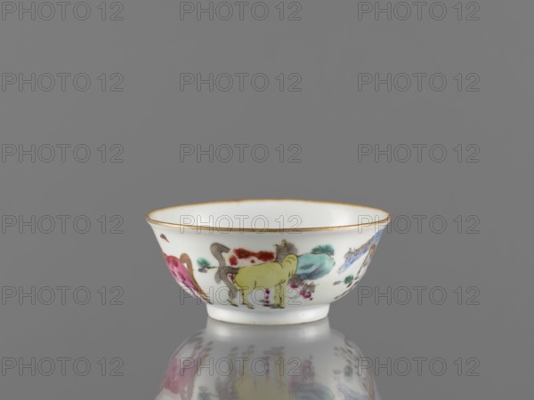 Bowl, colored porcelain, Qing 1644–1911, 19th century, China. Bowl on a ring-shaped foot, decorated on the outer wall with landscape motifs and horses. Vessel; height 5 cm, diameter 11.8 cm. Maker, inscription: Krieg doctor noted., A small ceramic bowl sits on a reflective surface against a plain gray background; the bowl is primarily white with a thin gold rim and painted decorations around its exterior showing a group of stylized animals in colors including yellow, pink, blue, green, brown, and red, with small green decorative accents and subtle shading; the reflection of the bowl and its painted motifs is visible on the surface beneath it.