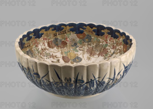 Bowl, Toyota Masanobu maker, signature on bottom in gold. Deep satsuma bowl on a low ring-shaped base, knobbed body with serrated rim resembling a chrysanthemum. Outer wall: clumps of blooming irises growing from water, flowers and leaves painted in two-tone blue enamel; stylized waves and ripples outlined in gold. Inner surface: large gold chrysanthemum rosette imperial coat of arms in the mirror with the Tokugawa family crest in the center; side wall filled with dense, colorful chrysanthemum flowers and butterflies painted in white, red, blue, gold, and green using overglaze enamels and gold. Top edge finished with a uniform cobalt blue valance ornament. Inscription reads: Satsuma yaki, Satsuma kuni, Ijun'in, Toyota Masanobu. 19th century Meiji period, Japan, Kagoshima. Ceramic vessel; height 12.0 cm, diameter 28.0 cm., A scalloped ceramic bowl shown against a neutral background with a pale exterior and deep blue painted floral motifs along the lower outer surface, a scalloped rim pa...