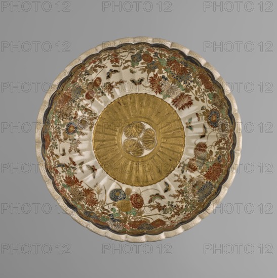Bowl by Toyota Masanobu author/craftsman. Deep bowl on a low, ring-shaped base with a knobbed body and serrated rim, the bowl reminiscent of a chrysanthemum flower. Outer wall decorated with clumps of blooming irises growing out of water encircling the bowl; flowers and leaves painted with two-tone blue enamel, outlines and stylized waves/ripples painted in gold. Inner surface decorated with a large gold chrysanthemum rosette imperial coat of arms in the mirror, with the Tokugawa family coat of arms inscribed in the center; side wall filled with a dense, colorful ornament of various chrysanthemum flowers and butterflies. The garden motif is painted in white, red, blue, gold, and green using overglaze enamel paints and gold, completed at the rim by a uniform cobalt blue valance ornament. Signature on the bottom painted in gold reading: Satsuma yaki, Satsuma kuni, Ijun'in, Toyota Masanobu. Satsuma ware, 2nd half of the 19th century Meiji period, Kagoshima, Japan. Vessel; height 12.0 cm, ...
