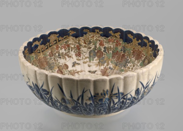 Deep bowl on a low ring-shaped base with a knobbed body and serrated rim, reminiscent of a chrysanthemum flower. Outer wall decorated with clumps of blooming irises growing from stylized water; flowers and leaves in two-tone blue enamel, outlines and wave/ripple motifs in gold. Inner surface bears a large gold chrysanthemum rosette imperial coat of arms in the mirror with the Tokugawa family coat of arms in the center; side wall filled with dense, colorful chrysanthemum flowers and butterflies in white, red, blue, gold, and green overglaze enamels and gold. Composition finished near the rim with a uniform cobalt-blue valance ornament. Signature painted in gold on the base reading: Satsuma yaki, Satsuma kuni, Ijun'in workshop, Toyota Masanobu. Satsuma ware, 2nd half of the 19th century Meiji period, Kagoshima, Japan. Vessel; height 12.0 cm, diameter 28.0 cm., A scalloped ceramic bowl sits against a neutral gray background, showing a cream exterior with a textured surface and a deep blue...