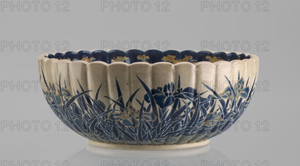 Bowl, Toyota Masanobu maker. Deep bowl on a low, ring-shaped base with a knobbed body and serrated rim, form reminiscent of a chrysanthemum flower. Outer wall decorated with clumps of blooming irises rising from water; flowers and leaves painted in two-tone blue enamel with gold outlines and stylized waves and ripples. Inner surface features a large gold chrysanthemum rosette imperial coat of arms in the mirror with the Tokugawa family coat of arms inscribed in the center; side wall filled with a dense, colorful ornament of various chrysanthemum flowers and butterflies. Painted in white, red, blue, gold, and green using overglaze enamels and gold; composition finished near the edge by a uniform cobalt blue valance ornament. Signature painted in gold on the bottom: Satsuma yaki, Satsuma kuni, Ijun'in, Toyota Masanobu. Satsuma ware, 19th century Meiji period, second half of the 19th century. Kagoshima, Japan. Ceramic vessel; height 12.0 cm, diameter 28.0 cm., A scalloped ceramic bowl sit...