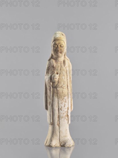 Figure of a standing woman — funerary figurine; creator, sculptor: Krieg doctor noted; images of women, figurines; Chinese art; Chinese culture;, repossession Jelenia Góra; female figurines; 10th century, China; sculpture; height 25.3 cm, A standing carved figure with a rounded head and a serene facial expression, wearing a long robe and a hooded head covering; the surface shows worn and weathered areas with patches of pale beige, off-white, light brown, and hints of muted pink, and the base reflects faintly on the smooth grey background.