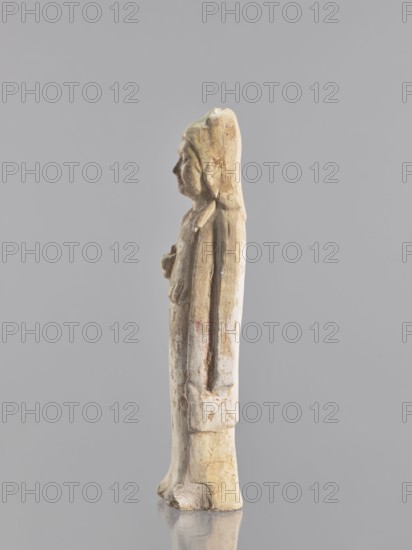 Figure of a standing woman — funerary figurine; creator, sculptor: Krieg doctor author; images of women; figurines; Chinese art; Chinese culture;, repossession Jelenia Góra; female figurines; 10th century ; China; sculpture; height 25.3 cm, A worn carved figure of a standing person shown in profile, with a long headdress or hood and a draped garment, rendered in pale beige and off-white tones with patches of light brown and faint reddish discoloration, standing on a rounded base with visible surface texture and small chips, set against a plain light gray background.