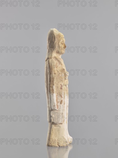 Figure of a standing woman - funerary figurine, creator, sculptor possibly Krieg doctor. Images of women, female figurines; Chinese art, Chinese culture; provenance: repossession Jelenia Góra; 10th century, China; sculpture, height 25.3 cm, A slender standing figure carved from a pale material with a weathered surface showing areas of beige, cream, off-white, and traces of dull pink and brown, shown in profile against a neutral gray background; the figure wears a long draped garment extending to the feet and a head covering that falls along the back, with subtle facial features, a straight posture, one arm close to the body and the other suggested by a raised area at the chest, and the base of the piece and its reflection visible on the smooth surface beneath.