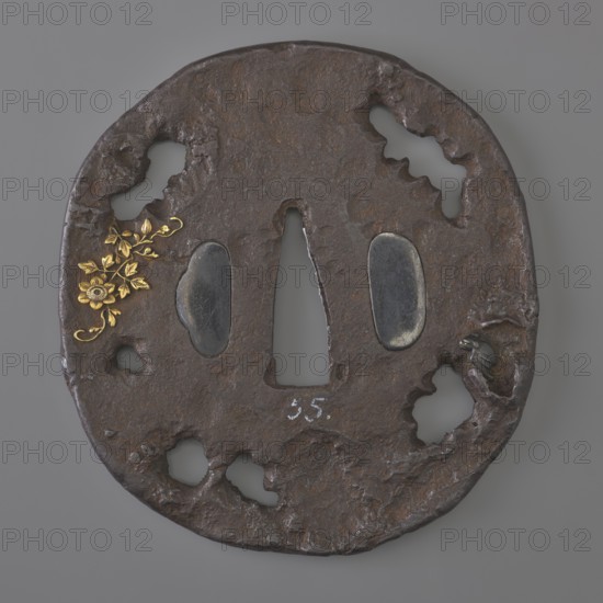 Crossguard tsuba, Oval tatemaru-gata tsuba with an irregular edge, surface textured to imitate rock and cut into openwork irregular elongated holes. Inlaid in high relief with a motif of a blooming rose bush, a snail and flying bats. Signed in flat gold inlay: italic character ju naga. 18th century Edo period, 1603–1868, Japan. Tsuba; height 8.0 cm, width 7.5 cm, depth 0.5 cm., A roughly circular dark brown metal object with an uneven, pitted surface and irregularly shaped cutouts around its perimeter and center, featuring a central vertical opening flanked by two smooth, oval dark gray insets; a decorative cluster of small gold-colored floral elements is attached on one side, and a small dark gray sculpted element resembling a tiny creature is set into a recess on the opposite side, with visible textures of rust and wear and overall colors including dark brown, dark gray, and gold.