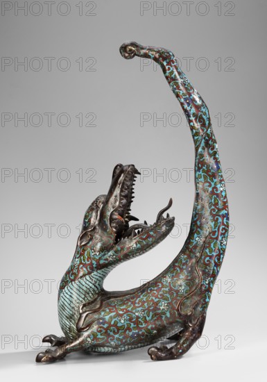 Dragon, gift, art of Southeast Asia, Southeast Asia culture, images of animals, fantastic creatures, dragons, 19th century, Southeast Asia, sculpture, height 51.0 cm, width 35.0 cm, A sculptural creature with an elongated curved body and upturned tail, shown in a three-quarter view, rendered in metal with intricate surface patterns in turquoise, blue, green, brown, and reddish tones; it has an open mouth with visible teeth and a forked tongue, a textured throat and ribbed underside, small rounded ears, clawed feet, and scrolling decorative motifs covering its surface against a plain light gray background.