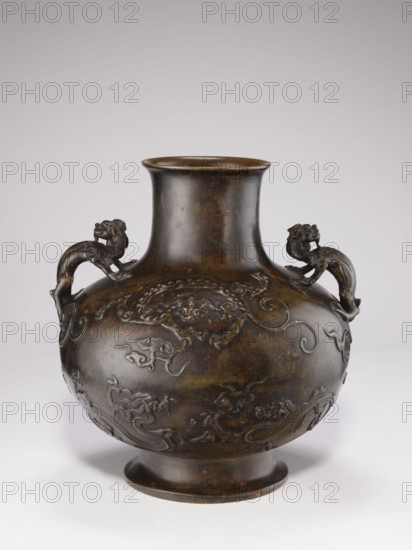 Vase with two dragon-shaped handles qiulong, from Ignacy Jan Paderewski 1860–1941. Chinese, Qing dynasty 1644–1911, 19th century. Porcelain vessel with dragons iconography and backdated signatures; belly in the shape of a flattened sphere; low, round foot; cylindrical short neck with a very slightly flared spout. Height 38 cm, diameter 15 cm, weight 10.0 kg., A rounded metal vase with a narrow neck and flared rim sits against a pale background; the surface is dark brown with areas of lighter brown and subtle greenish tones, and the body features raised decorative motifs in the same metal tone. A pair of sculpted handles shaped like stylized animal figures rise from the shoulder of the vase, matching the dark brown coloration. The decoration includes flowing, swirling relief patterns wrapping around the rounded midsection and lower body, all rendered in the same metallic hues. The base flares slightly and shares the dark brown finish, and the overall surface shows a patinated, aged appe...