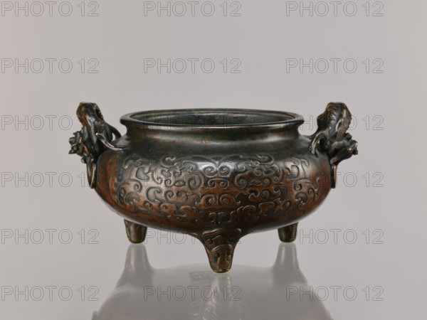 Ding incense burner, Chinese, Qing 1644–1911, 19th century. Based on ceramics from the Xuande era 1426–1435 of the Ming dynasty. Ceramic vessel with dragon-shaped handles; decorations include lizards, dragons, and taotie motifs; backdated signatures. Height 13.1 cm; diameter 21.4 cm., A dark bronze vessel with a rounded body sits on a smooth reflective surface, supported by three short legs and flanked by sculpted handles resembling stylized animal heads; the body is decorated all around with raised swirling cloud-like patterns. The surface shows variations of dark brown and deep greenish-black patina with subtle areas of warmer brown and hints of reddish bronze; the handles have slightly darker tones and small highlights where the metal catches light. The background is a neutral pale gray, and the reflection of the vessel is visible beneath it.
