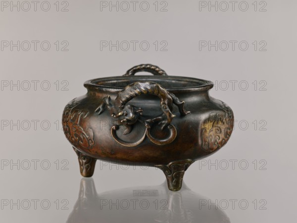 Ding incense burner, Chinese, Qing 1644–1911, backdated signatures, dragon-shaped handles. Based on ceramics from the Xuande era 1426–1435 of the Ming dynasty; 19th century, China. Ceremonial/ritual vessel with lizards, dragons, and taotie iconography. Height 13.1 cm; diameter 21.4 cm., A low, rounded tripod censer with a wide mouth and a curled handle on top, cast in a dark metallic material with polished highlights in brown, bronze, gold, and greenish patina; the body rests on three short curved legs and bears raised decorative motifs, while a sculpted mythical creature with a sinuous body and curled tail clings to the front edge holding a ring in its mouth, and the object sits on a reflective pale surface against a neutral light gray background.