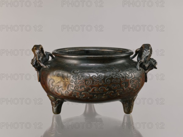 Ding incense burner, Chinese, Qing 1644–1911, 19th century. Based on Xuan De era 1426–1435 Ming ceramics. Bronze vessel incense burner with dragon-shaped handles; taotie and dragon iconography, lizard motifs. Backdated signatures. Height 13.1 cm, diameter 21.4 cm. Ceremonial/ritual vessel., A low, rounded metal vessel with a wide rim and a bulbous body stands on three short curved legs; the body is decorated with swirling raised patterns and has sculpted handles on opposite sides shaped like stylized animal heads with curled features. The surface shows a mix of dark bronze, brown, and subtle greenish patina with areas of warmer golden-brown highlights and darker almost black shadows. The vessel sits on a reflective surface and is set against a neutral light gray background.