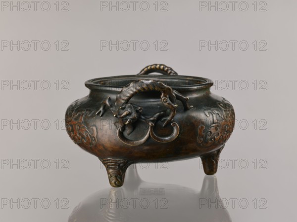 Ding incense burner, Chinese; Qing 1644–1911, 19th century, based on ceramics from the Xuande era 1426–1435 during the Ming dynasty. Bronze vessel, ceremonial/ritual incense burner with dragon-shaped handles; motifs include lizards, dragons, taotie; backdated signatures. Height 13.1 cm, diameter 21.4 cm., A rounded metal vessel with a dark brown and greenish patina rests on a reflective surface, supported by three short ornate legs; the body shows raised swirling decorative motifs and sculpted handles shaped like a curled animal with a textured body gripping circular rings, and a braided loop handle spans the rim; visible colors include dark brown, greenish bronze, and hints of golden wear.