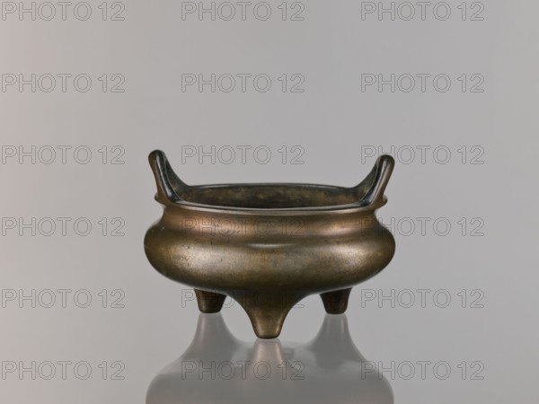 Li-type incense burner, ceremonial/ritual Li-type vessel; Chinese, Qing dynasty 1644–1911, 18th century; round, flattened incense burner on three low legs; backdated signatures; height 10.0 cm, diameter 15.7 cm., A small round three-legged bronze vessel with a bulbous body and a flared rim, showing a patina of dark brown, greenish bronze, and small areas of lighter golden bronze; the rim has two upward-curving handles opposite each other, and the vessel sits on a reflective surface that mirrors its shape.
