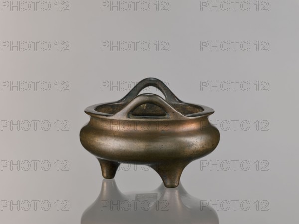 Li-type incense burner, Chinese, Qing dynasty 1644–1911, 18th century. Round, flattened Li-type ritual/ceremonial incense burner on three low legs, with backdated signatures. Height 10.0 cm, diameter 15.7 cm., A small rounded metal vessel with a smooth, slightly bulbous body and three short tapering legs, shown against a plain light gray background; the vessel has an open top with an integrated looped handle structure that rises above the rim in a double-arched shape, and the metal surface shows a patina with shades of bronze, brown, muted gold, and hints of darker gray, with subtle areas of wear and slight greenish spots; the vessel casts a soft reflection on the glossy surface beneath it.