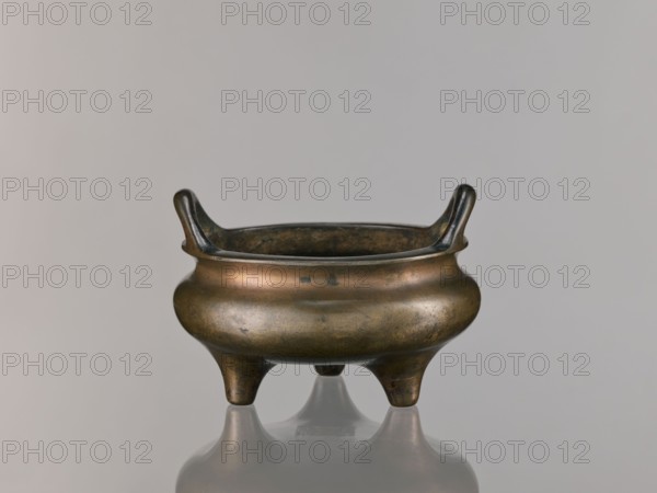 Li-type incense burner, Qing dynasty 1644–1911, 18th century, Chinese, round flattened form on three low legs, Li-type/ritual/ceremonial vessel, incense burner, with backdated signatures, height 10.0 cm, diameter 15.7 cm, A small metal vessel with a rounded body and a wide rim sits on a reflective surface, supported by a group of three short, tapered legs; the rim rises into a pair of upward-curving handles on opposite sides. The vessel shows a patina with shades of brown, bronze, dark greenish spots, and darker brown to almost black areas inside the rim and on the handles. The background is a smooth, even light gray, and the reflection beneath the vessel reproduces its colors and shape faintly.