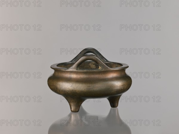 Li-type incense burner, Chinese, Qing dynasty 1644–1911, 18th century. Round, flattened Li-type ritual/ceremonial incense burner on three low legs, with backdated signatures. Height 10.0 cm; diameter 15.7 cm., A small metal vessel with a rounded body and a flared rim stands on three short tapered legs; it has a pair of curved handles that arch above the rim and mirror each other, and the metal surface shows a patina with tones of brown, bronze, dark green, and hints of black, set against a smooth light gray background and reflected faintly on the glossy surface beneath.