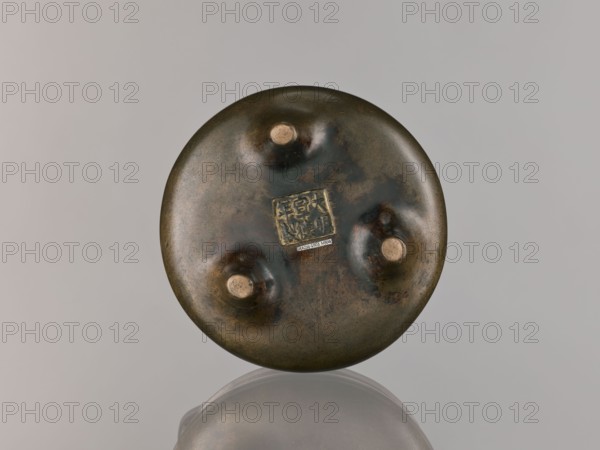 Li-type incense burner, Chinese; Qing dynasty 1644–1911, 18th century; round, flattened incense burner on three low legs; Li-type ceremonial/ritual vessel; backdated signatures; height 10.0 cm, diameter 15.7 cm., A round, slightly convex object with a mottled dark bronze surface showing areas of brown, green, and black, with three raised circular bumps that have lighter tan centers, and a small square embossed detail near the center; the object sits against a neutral pale gray background and its underside is reflected faintly below, showing similar bronze and brown tones.