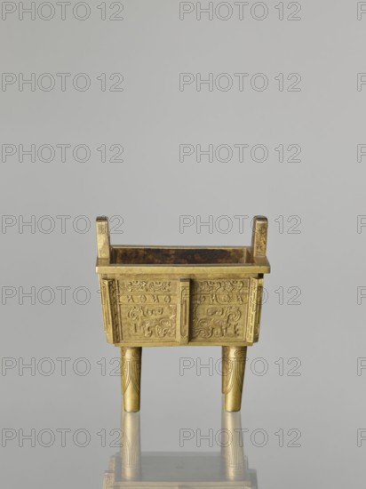 Fangding incense burner with cover, Chinese, Qing dynasty 1644–1911, 19th century. Bronze vessel decorated on the walls with archaic ornament in flat relief, taotie and fantastic creature motifs; ceremonial vessel. Height 15.2 cm, width 14.0 cm, depth 10.9 cm., A small rectangular metal vessel with a golden color sits on three tapered legs and has two short upright handles at opposite ends; the vessel's sides feature raised decorative patterns with curving motifs and panels, and the legs show incised linear designs; the interior appears darker with a rich brown tone, and the object casts a subtle reflection on a smooth gray surface beneath it against a pale gray background.
